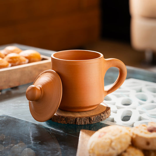 Terracotta mug with lid on a wooden coaster, surrounded by pastries on a marble surface.