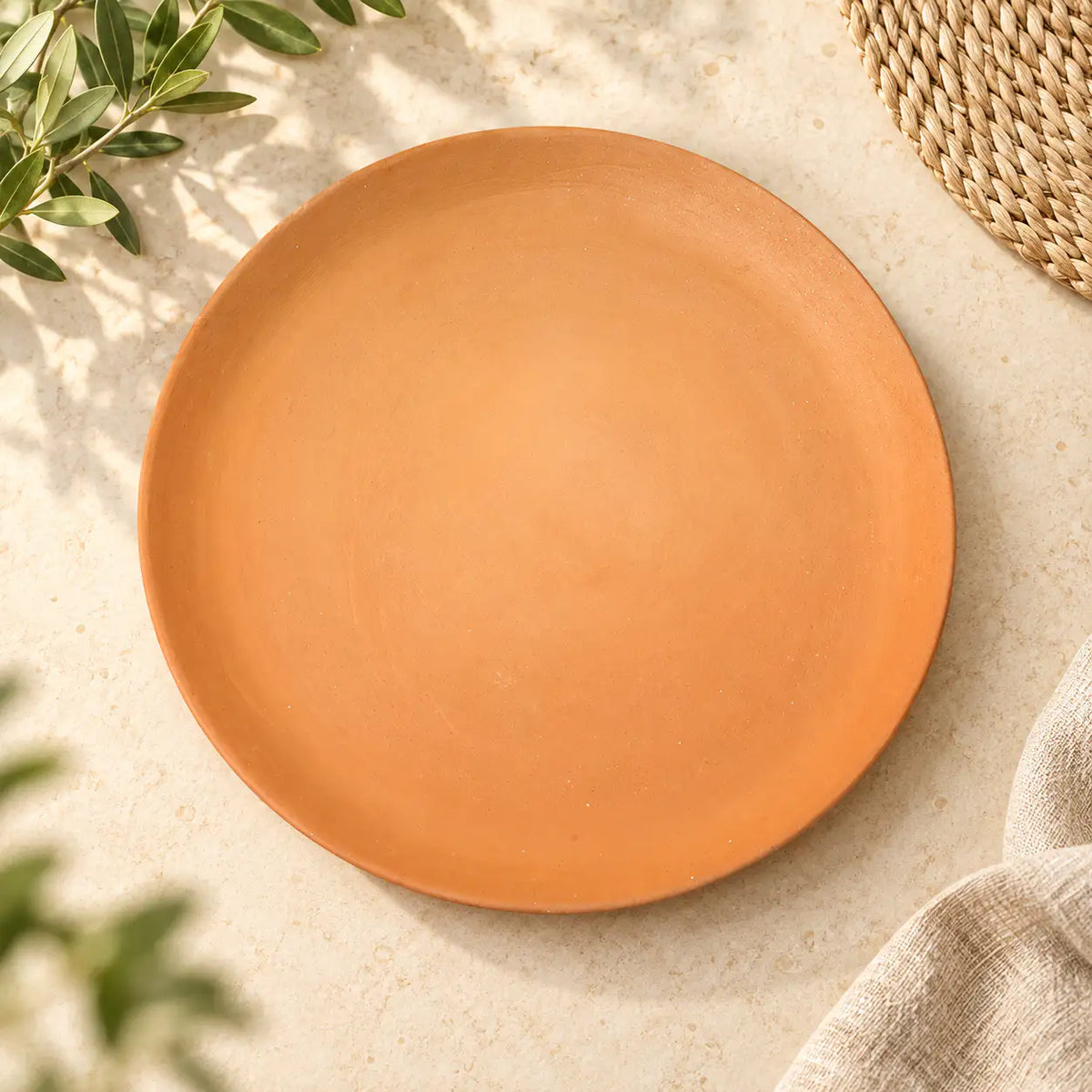 A round terracotta plate with a matte finish, photographed from a top-down angle on a textured beige surface with decorative olive branches and a woven mat.