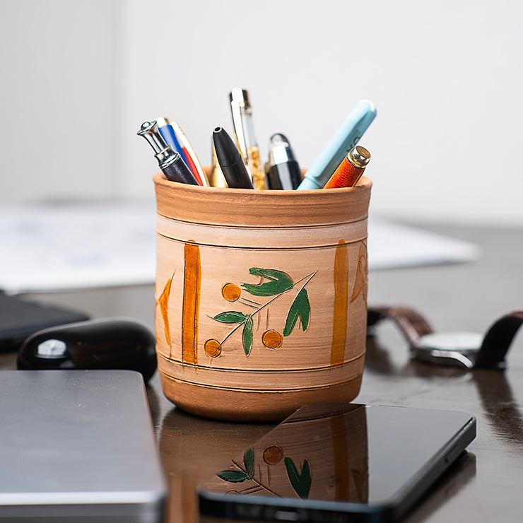 ClayNest handmade terracotta pen holder in cylindrical shape with hand-painted leaf motif, holding pens on a desk