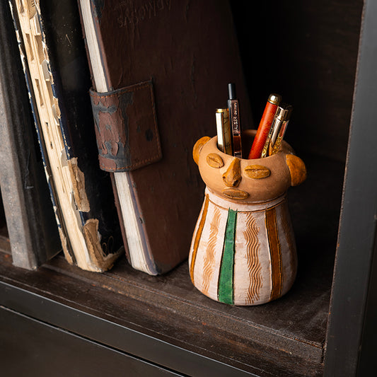 ClayNest handmade terracotta pen stand with sculpted tribal face design and hand-painted details, holding pens on a bookshelf