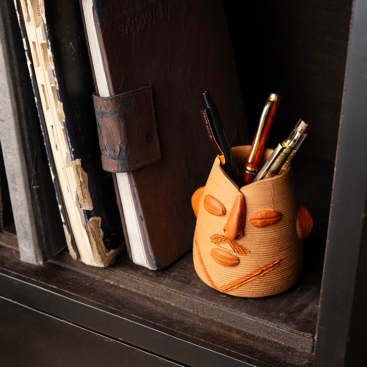 ClayNest handmade terracotta pen stand with sculpted face design, textured finish, holding pens on a bookshelf
