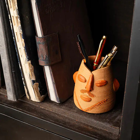 ClayNest handmade terracotta pen stand with sculpted face design, textured finish, holding pens on a bookshelf