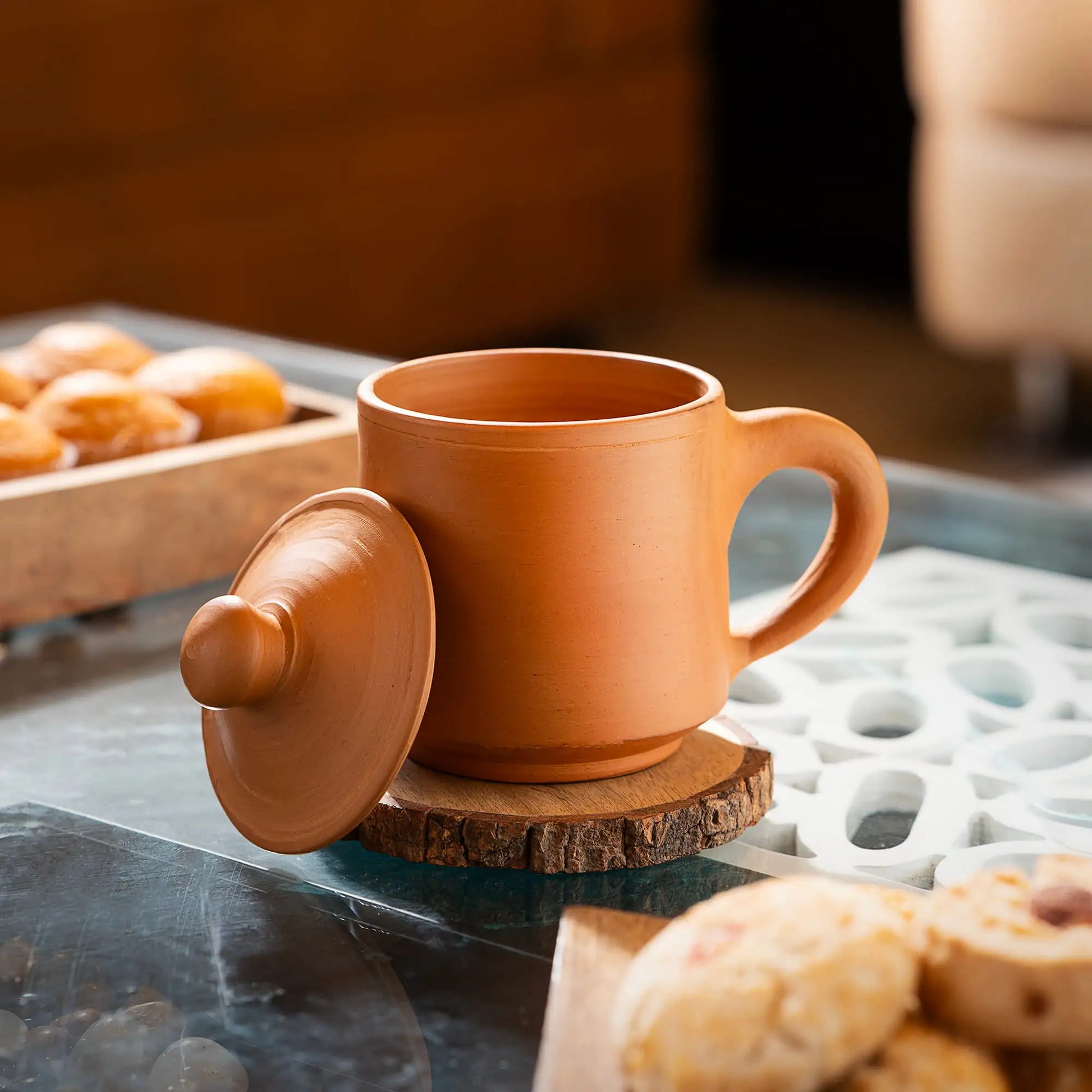 Terracotta mug with lid on a wooden coaster, surrounded by pastries on a marble surface.