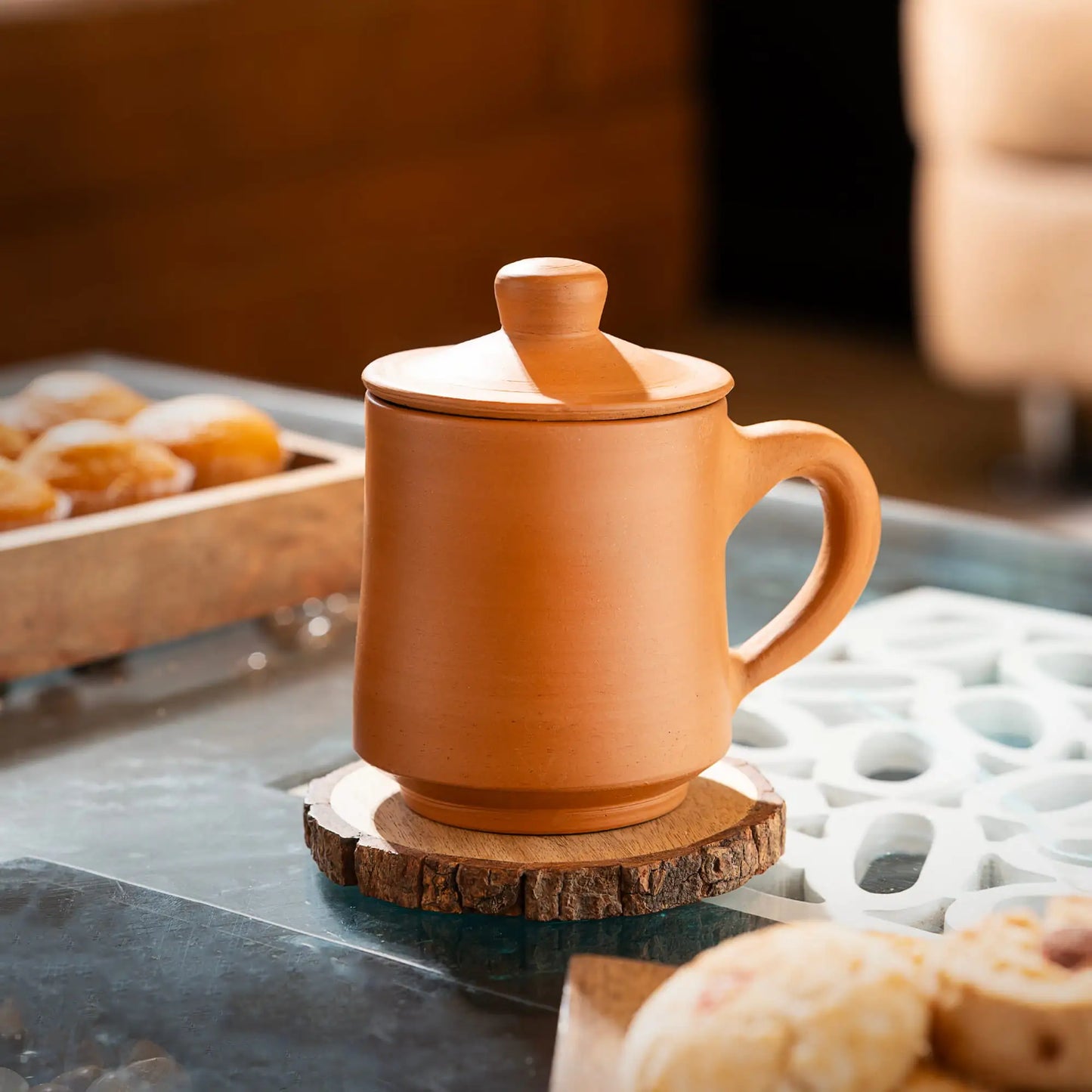 Terracotta mug with lid on a wooden coaster, surrounded by pastries.