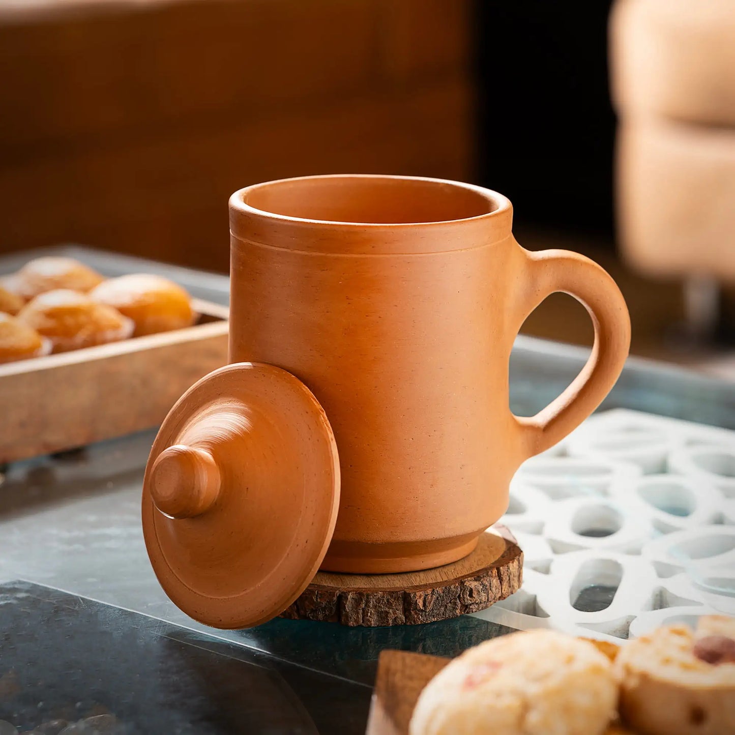 Terracotta mug with lid on a wooden coaster, surrounded by pastries on a table.