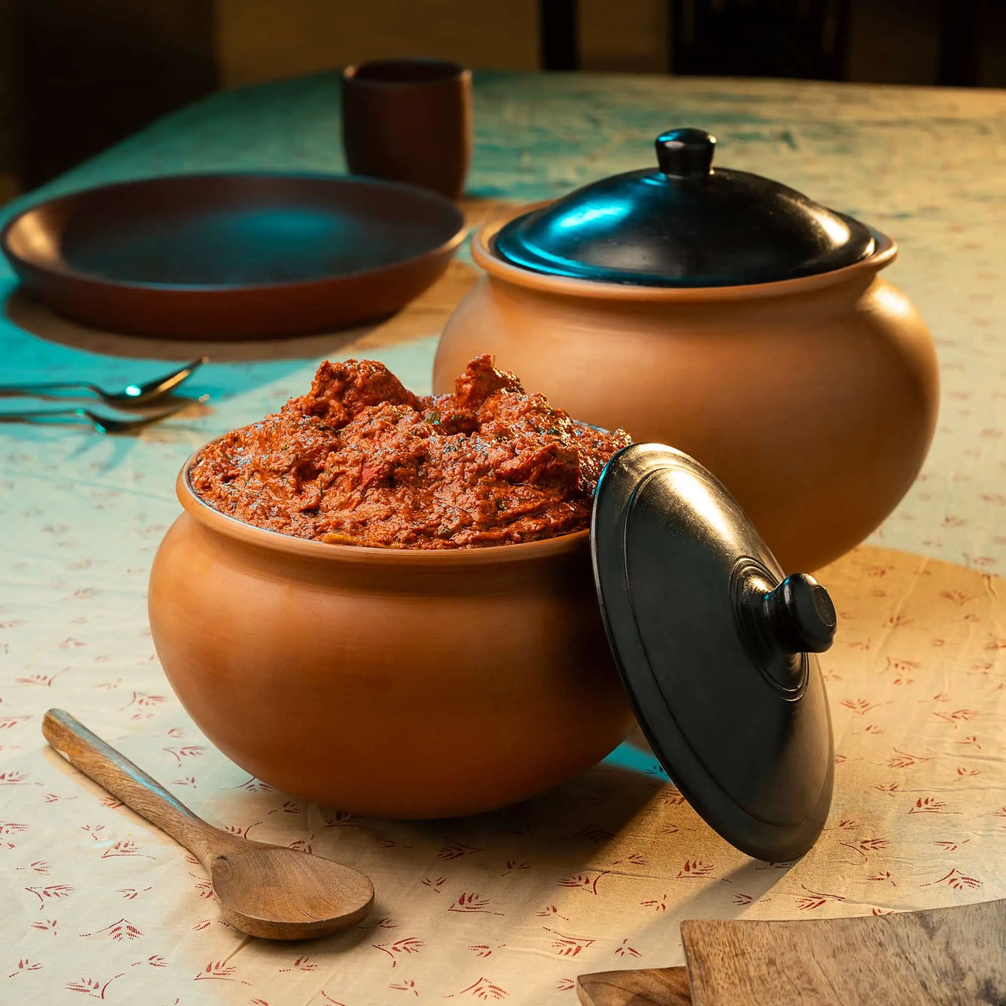 Two terracotta pots with lids on a table with a wooden spoon and a dish in the background.