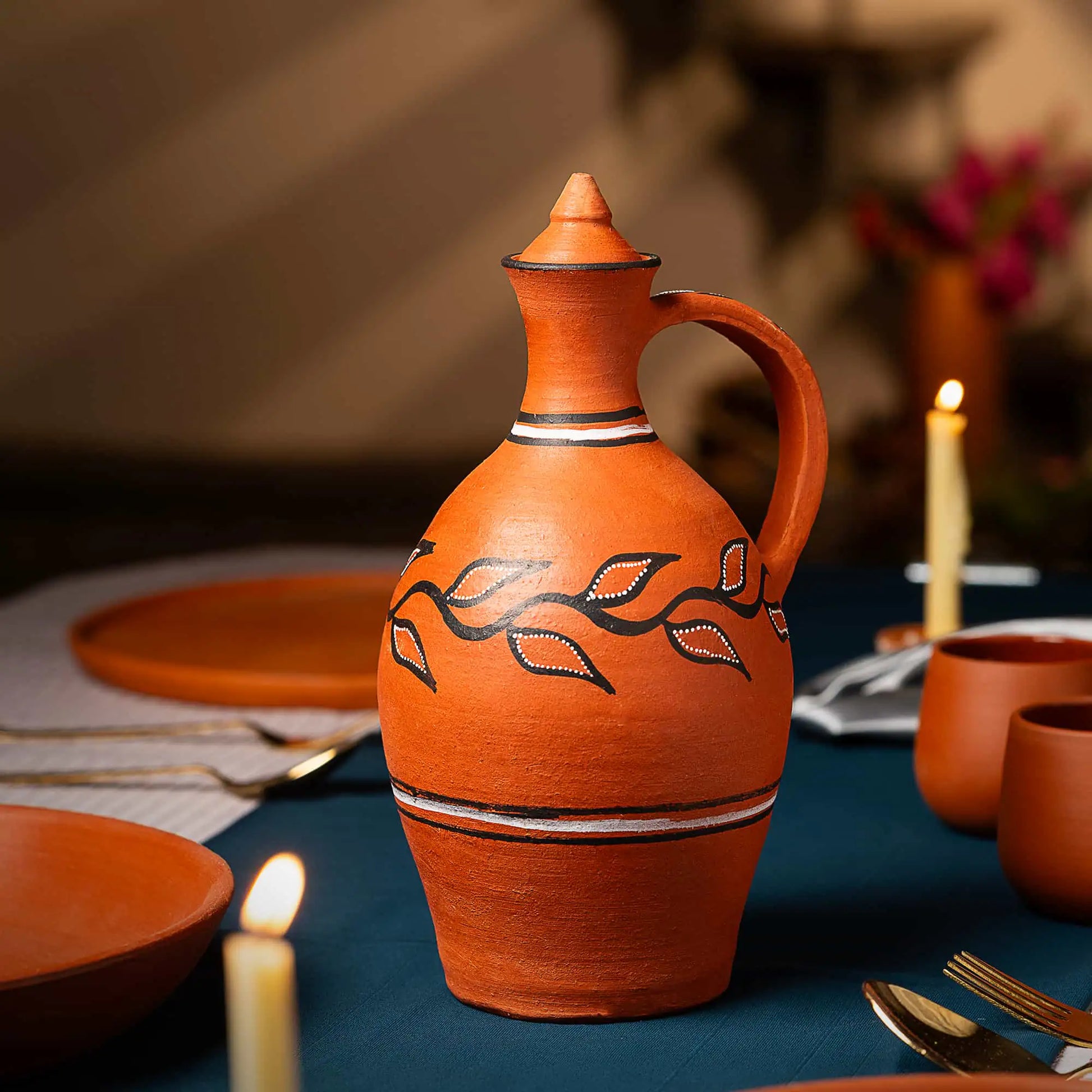 Terracotta pitcher with decorative elements on a table setting with candles and plates.