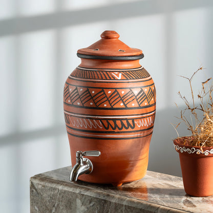 Terracotta jar with decorative patterns and a silver faucet on a marble surface.