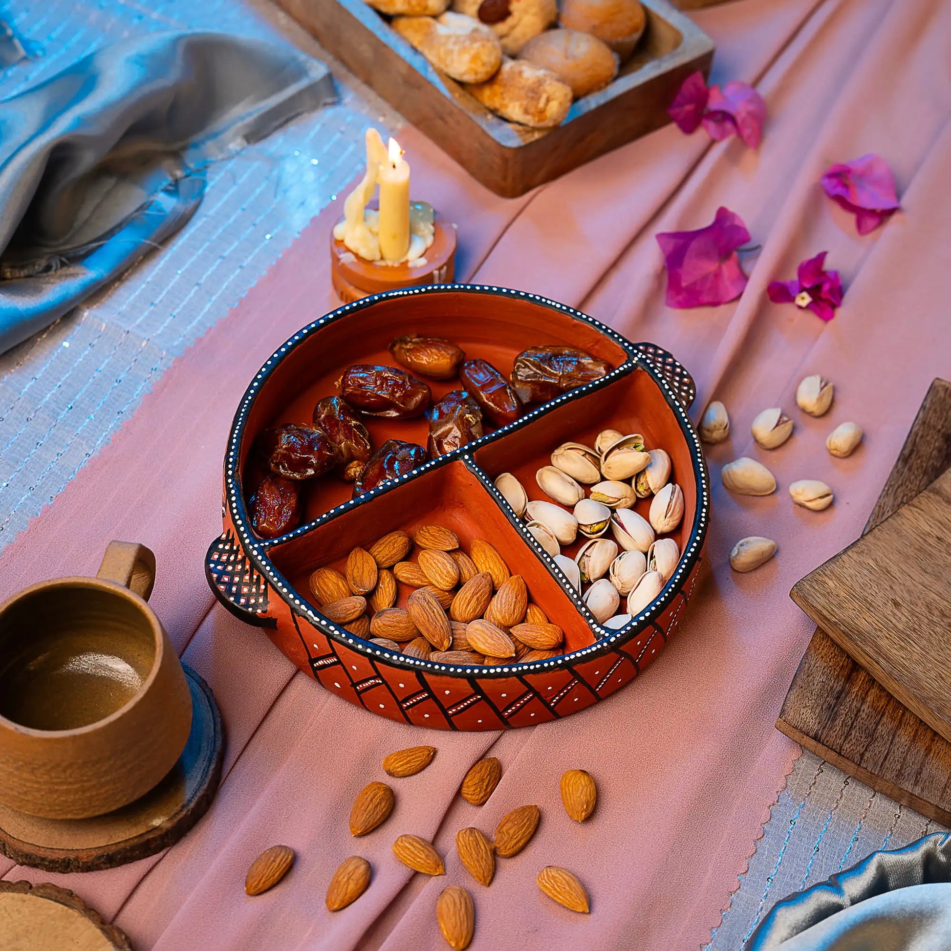 Dried fruits and nuts in a divided Terracotta bowl on a pink fabric with a candle and cup.