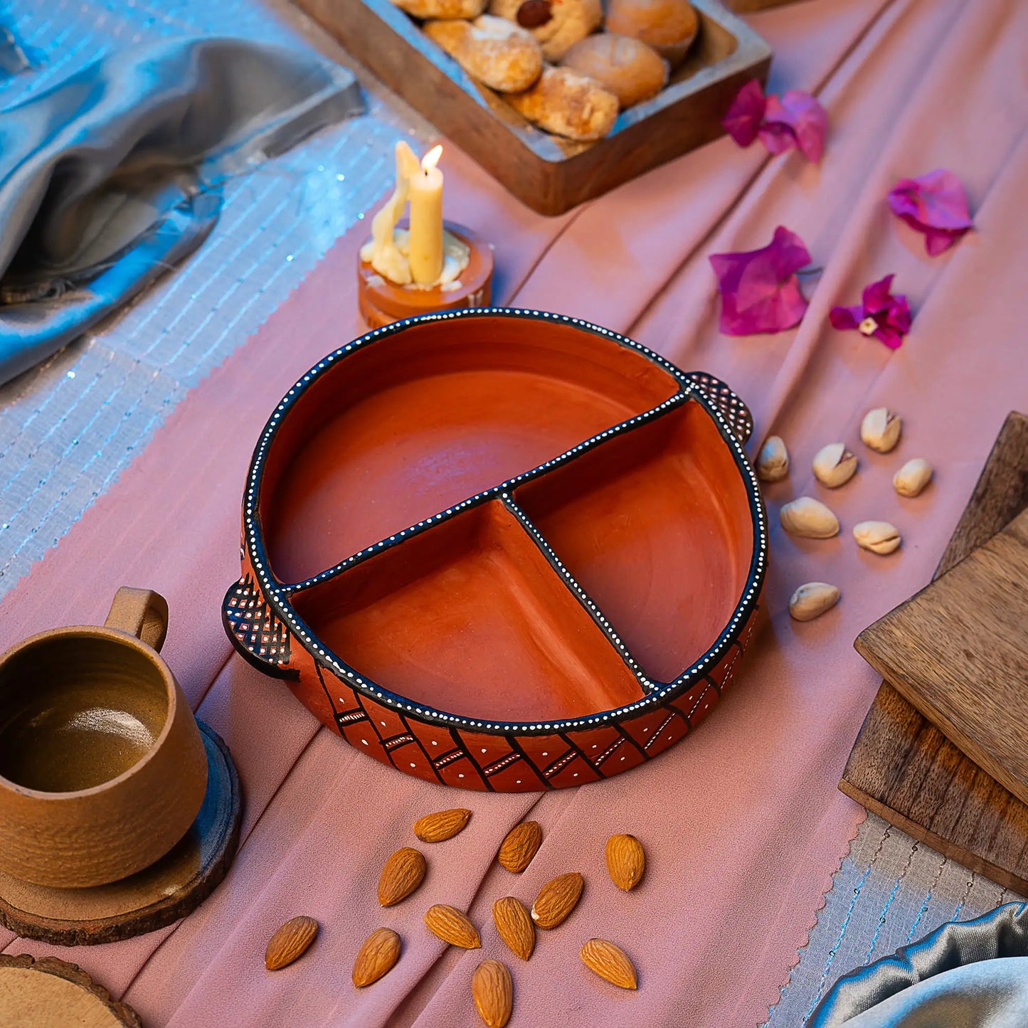 Terracotta dish with a cup on a pink fabric surface with nuts and flowers.