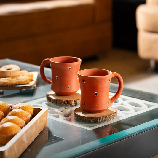 Two terracotta mugs with decorative holes on a glass table with pastries.