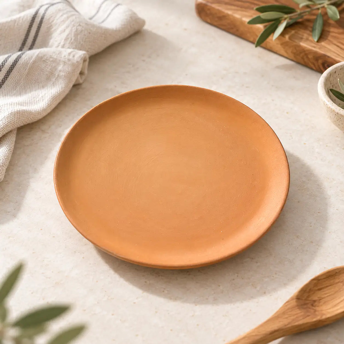 A round, matte terracotta plate is displayed on a light beige textured surface, surrounded by rustic kitchen props including a wooden spoon, a striped cloth napkin, and green leaves.