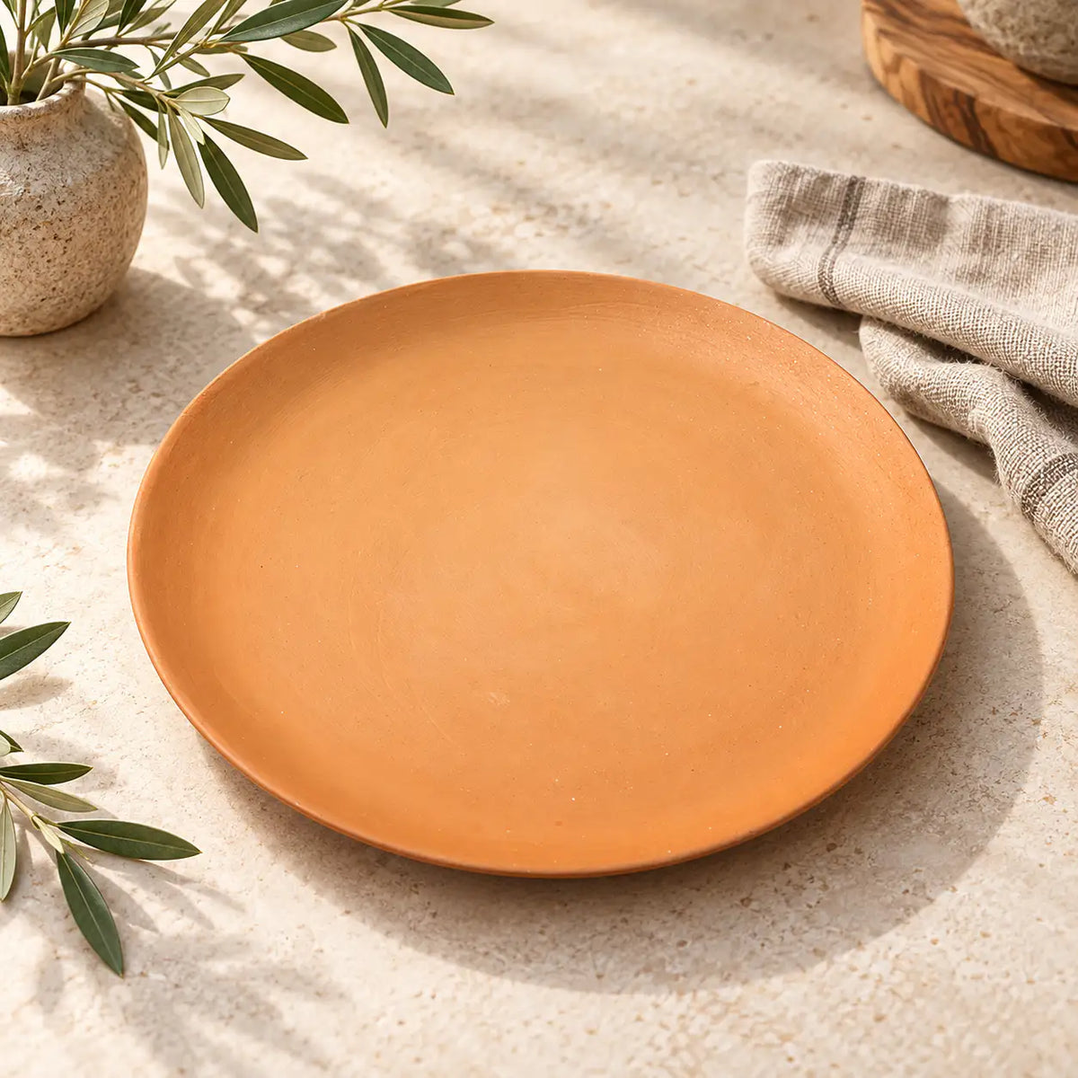 A round terracotta plate with a matte finish, styled on a textured beige stone surface with olive branches, a striped linen napkin, and a wooden board in the background.