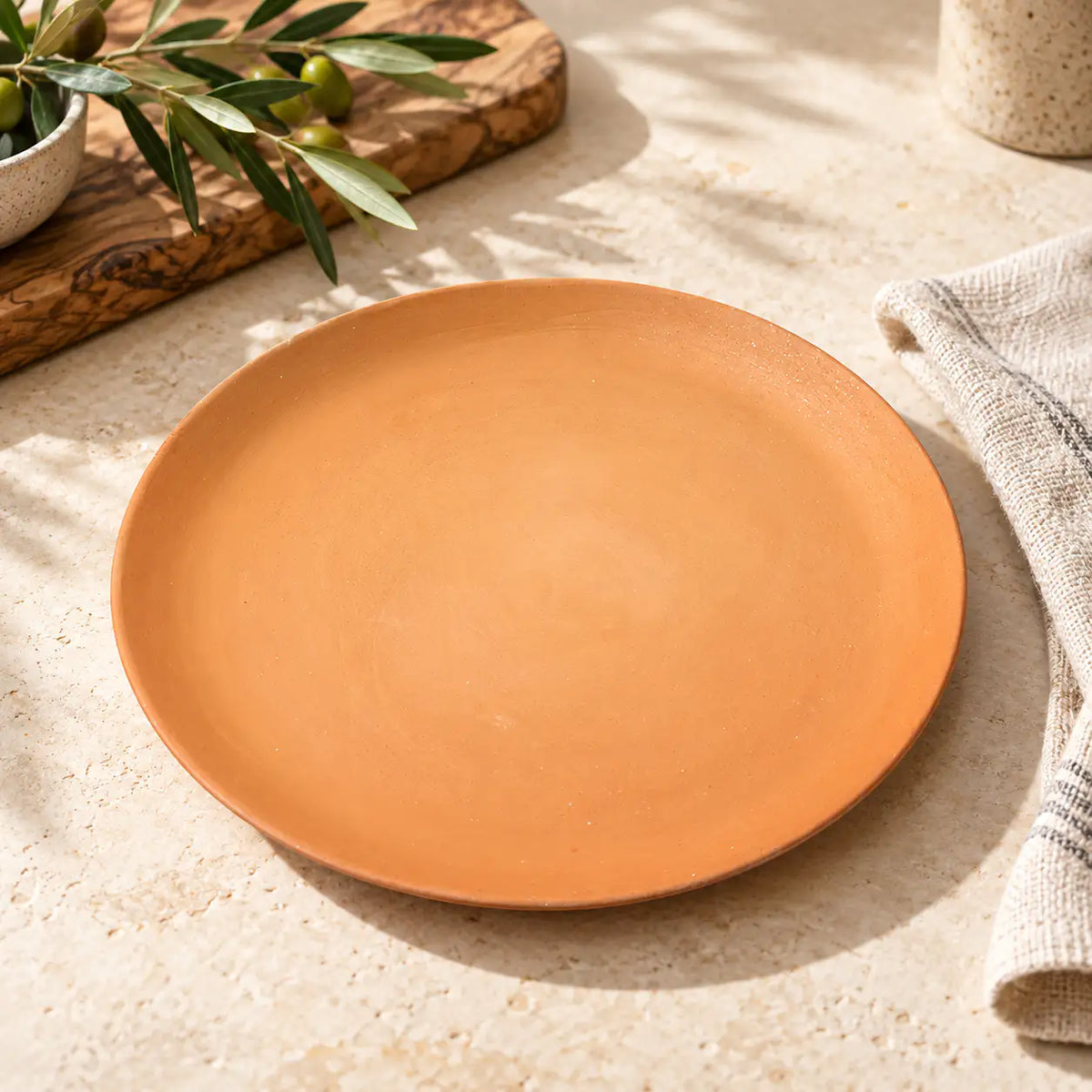 A round terracotta plate with a matte finish, styled on a textured beige stone surface with natural shadows, olive branches, and a linen napkin.