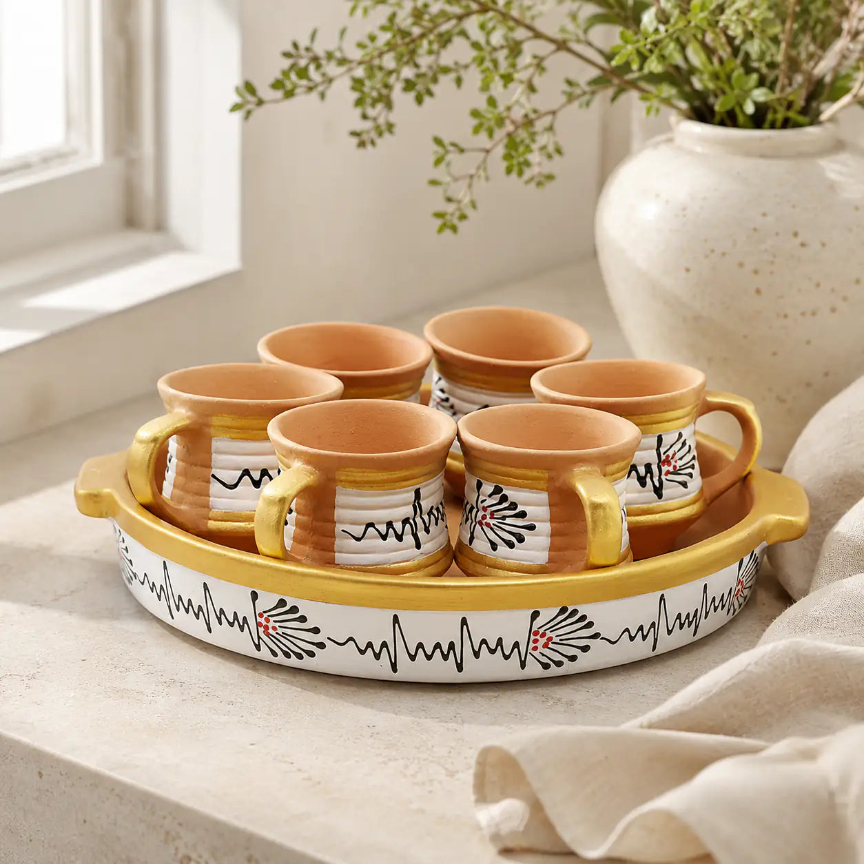 Set of clay mugs with a decorative tray on a light surface with a plant in the background.