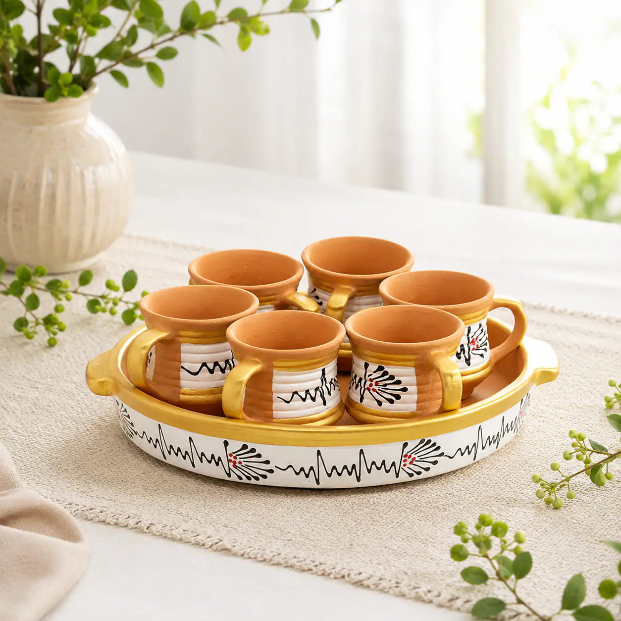 Set of terracotta mugs with decorative handles on a matching tray, placed on a light surface with greenery.